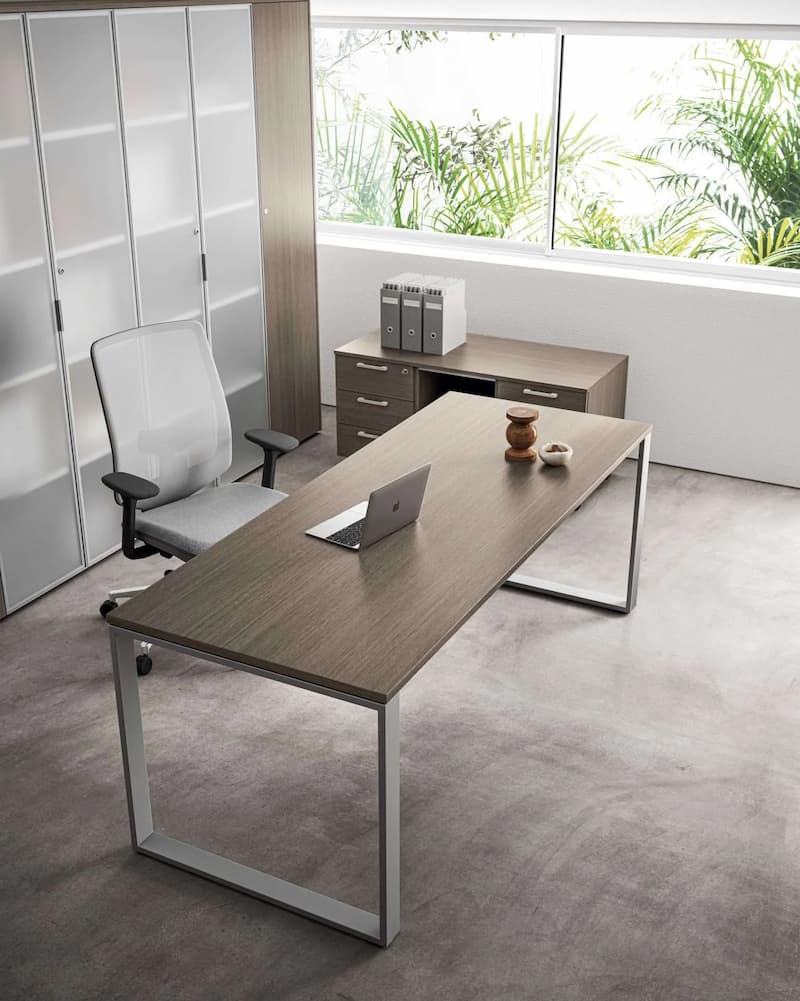 L'immagine mostra un ufficio moderno con una scrivania in legno, un laptop e oggetti decorativi. Una sedia ergonomica è davanti a un mobile con raccoglitori. Grandi finestre offrono vista su piante verdi, creando un'atmosfera luminosa e tranquilla.